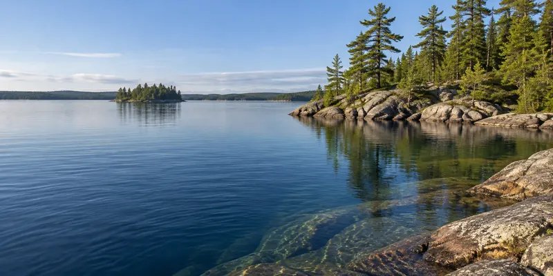 Clear lake water around a low granite point with tall white pines and a distant island.